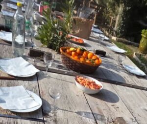 Table en bois rustique dressée pour un repas en plein air au Mas de la Sacristière, avec une bouteille d'eau, des verres, un panier de mandarines fraîches et un bol de crevettes, sous le soleil doux de Provence pour le Premier Janvier.