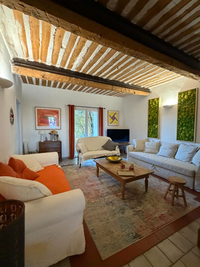Salon chaleureux et lumineux d'un mas provençal, photographié depuis un angle légèrement oblique. Le plafond est caractérisé par des poutres apparentes en bois foncé et des solives peintes en blanc et bois clair. Au premier plan à gauche, un grand canapé blanc à housse avec des coussins orange vif. Au centre, une table basse rectangulaire en bois clair repose sur un grand tapis aux motifs géométriques et couleurs douces. À droite, un autre grand canapé blanc. Au fond, une troisième banquette blanche fait face à une télévision noire. Deux grands panneaux muraux vert vif, composés de mousse végétale stabilisée, encadrent une applique murale sur le mur du fond. Des fenêtres avec rideaux rouges laissent entrevoir la verdure extérieure. Le sol alterne entre des carreaux de terre cuite et le tapis.