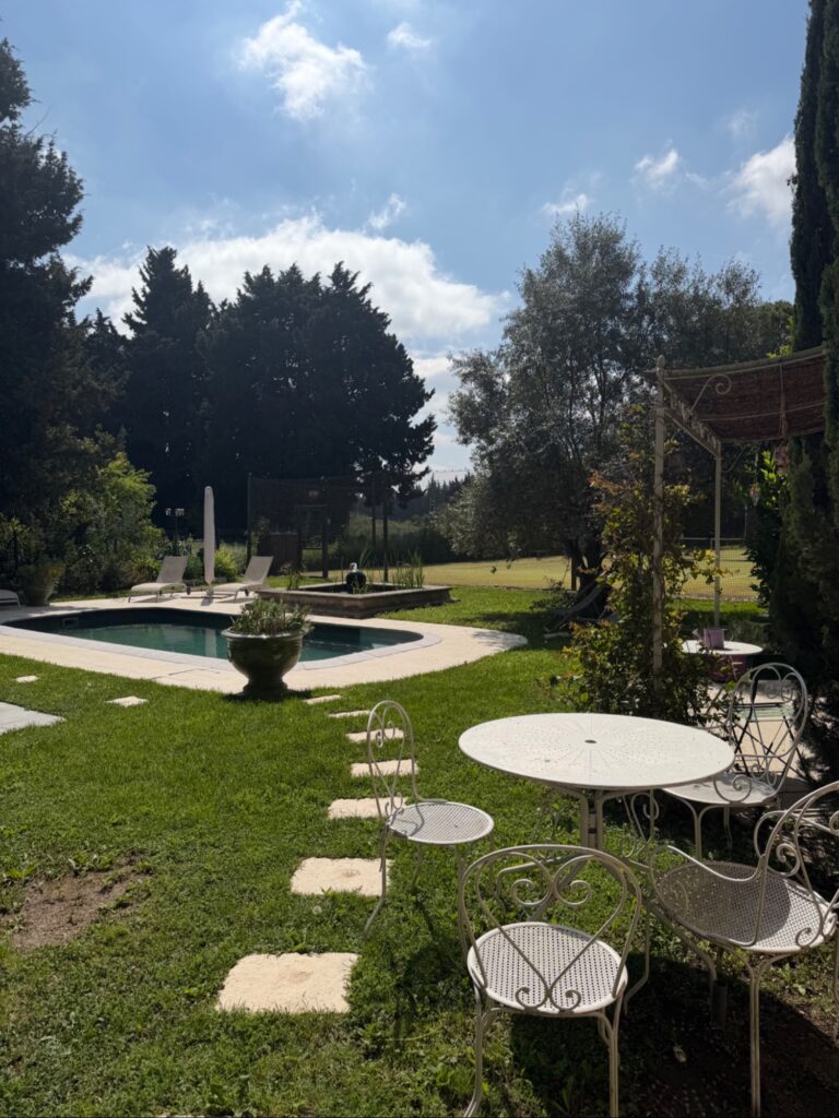 Vue panoramique sur la piscine limpide et le jardin verdoyant du Mas de la Sacristière à Jonquerettes, avec une table et des chaises en fer forgé, sous un ciel bleu d'été provençal.