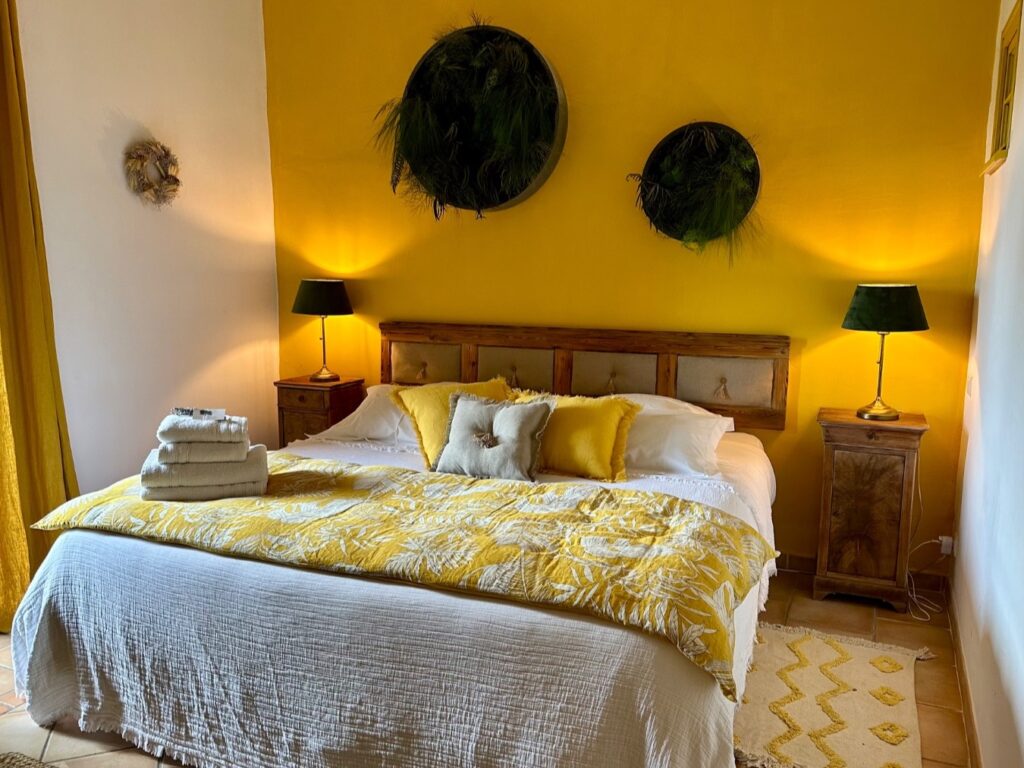 Chambre d'hôtel double lumineuse et chaleureuse avec murs jaunes, lit blanc et jeté de lit jaune, tables de chevet en bois et décorations végétales, évoquant le soleil de Provence.