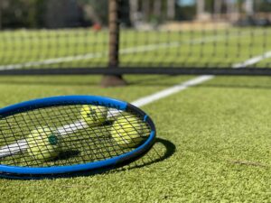 Raquette de tennis bleue et balles sur un court en gazon synthétique, avec filet en arrière-plan, sous le soleil de Provence. Représente l'option sport et activité physique proposée.
