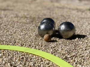Trois boules de pétanque Obut et un cochonnet sur un terrain de gravier ensoleillé, avec un marqueur jaune, illustrant une activité sportive provençale.