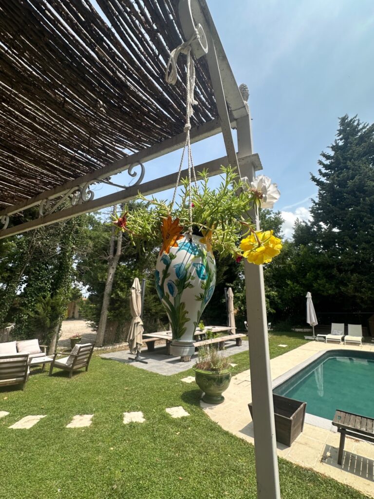 Jardin ensoleillé d'un mas provençal avec une pergola ombragée, un grand vase suspendu fleuri, un salon de jardin confortable, une piscine bleue et des arbres méditerranéens, idéal pour la détente et les vacances.
