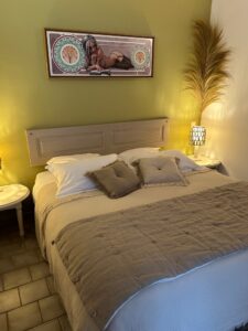 Chambre double douillette et lumineuse au Mas de la Sacristière, avec un grand lit double, une tête de lit en bois clair, une literie douce aux tons naturels (blanc et beige), des tables de chevet avec lampes, et une œuvre d'art murale distinctive.