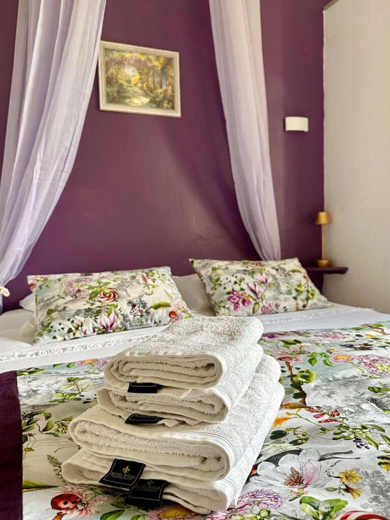 Lit double avec parure florale et serviettes blanches pliées dans une chambre d'hôtes aux murs violets, sous un voile de lit, évoquant la douceur de l'été au Mas de la Sacristière.
