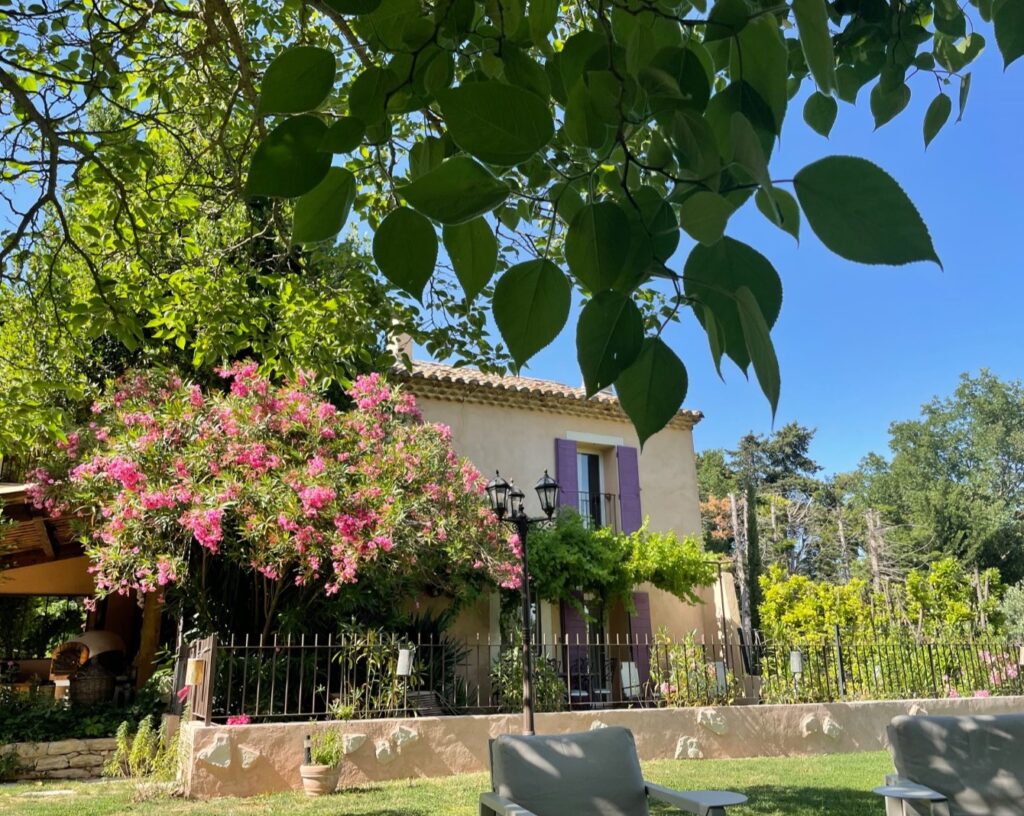 Vue d'un mas provençal traditionnel avec des volets violets, un jardin luxuriant et un grand oléandre rose en pleine floraison sous le soleil de juin.