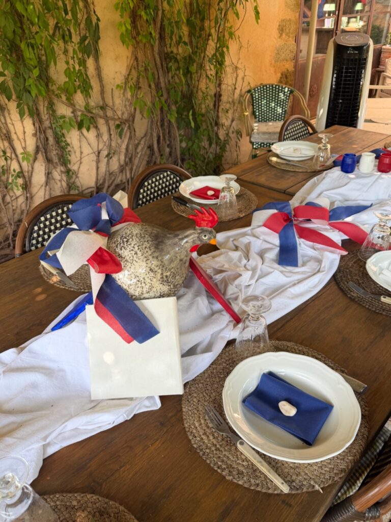 Table en bois décorée aux couleurs de la France avec un coq en céramique pour le 14 Juillet au Mas de la Sacristière en Provence, ambiance festive en extérieur.
